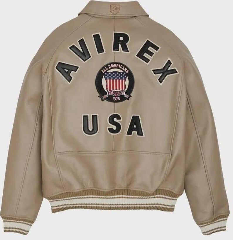 May include: A tan leather bomber jacket featuring a fold-down collar with a small logo patch. The back displays large black applique lettering reading AVIREX and USA, with a central circular patch showing the American flag and text 1975.