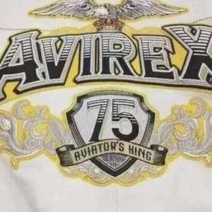 May include: A white jacket featuring a large embroidered graphic on the back. The design includes an eagle, the text AVIREX in black and yellow, a shield with the number 75, and a banner with the text AVIATOR'S KING, all in yellow and grey.