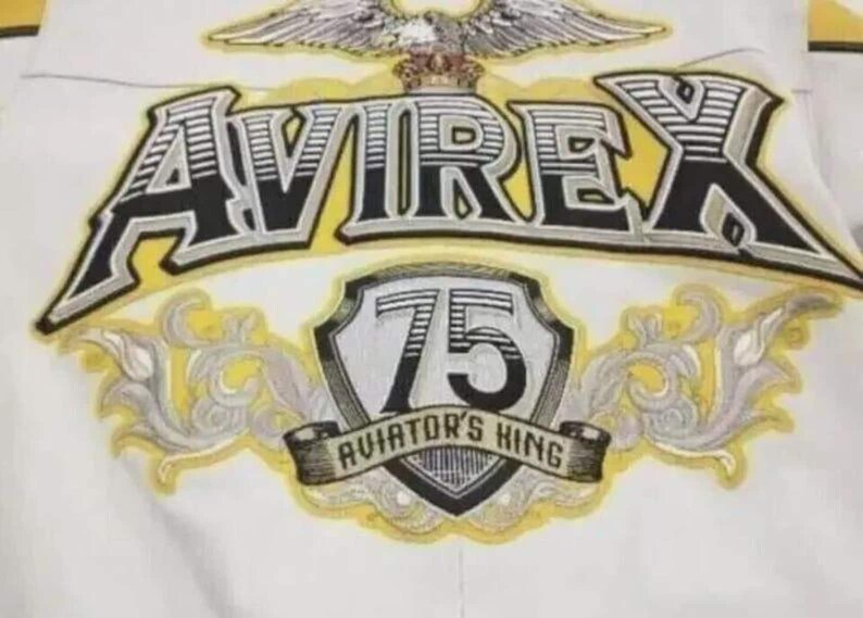 May include: A white jacket featuring a large embroidered graphic on the back. The design includes an eagle, the text AVIREX in black and yellow, a shield with the number 75, and a banner with the text AVIATOR'S KING, all in yellow and grey.