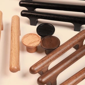 May include: A collection of cabinet hardware including long bar pulls and round knobs made from light oak, dark walnut, and black finished wood. The assortment features various lengths and finishes, showcasing natural wood grain textures and smooth surfaces.