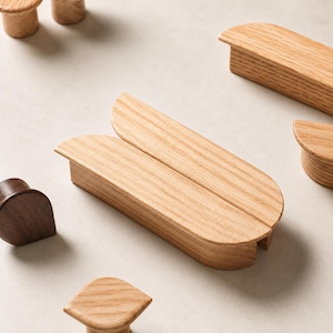 Puede incluir: Una colección de herrajes de madera para muebles que incluye varios tiradores de roble de tono claro con un diseño liso, alargado y redondeado, junto con un pomo de nogal de tono oscuro y otros pequeños tiradores de madera con veta visible.