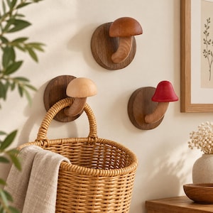 Può includere: Tre ganci da parete a forma di fungo, ciascuno con una base circolare in legno scuro e un gambo. I cappelli dei funghi sono rifiniti in legno chiaro, marrone medio e rosso. Un cesto intrecciato è appeso al gancio con il fungo chiaro.