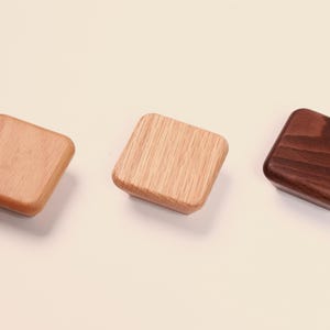 May include: Three square wooden knobs with rounded corners, arranged in a horizontal row. The left knob is light tan, the center knob features a visible light wood grain pattern, and the right knob is a dark brown wood with a smooth finish.