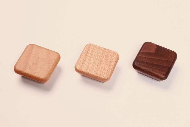 May include: Three square wooden knobs with rounded corners, arranged in a horizontal row. The left knob is light tan, the center knob features a visible light wood grain pattern, and the right knob is a dark brown wood with a smooth finish.