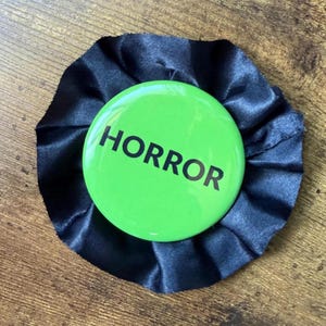 May include: A circular lime green pin-back button featuring the word HORROR printed in bold black capital letters. The button is centered on a black fabric rosette with gathered, pleated edges, creating a layered, circular badge design.