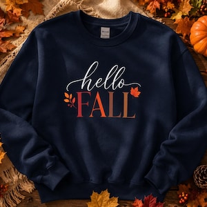 May include: A navy blue crewneck sweatshirt featuring the white cursive text hello above the word FALL in a gradient of orange and red. The design includes a small orange leaf graphic and a sprig of orange berries on the left side of the text.