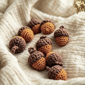 May include: A collection of small, handmade crochet acorns featuring dark brown caps and light tan, textured bodies. Each individual acorn is crafted with yarn, showcasing a rounded shape and a small loop at the top, arranged in a cluster.