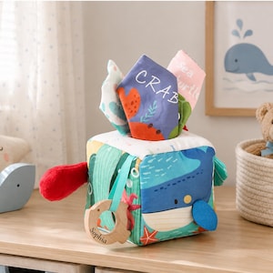 May include: A soft fabric sensory cube featuring colorful ocean-themed illustrations of a blue whale, starfish, and coral. The cube has protruding fabric tags labeled CRAB and SEA TURTLE, and a wooden ring engraved with the name Sophia attached.