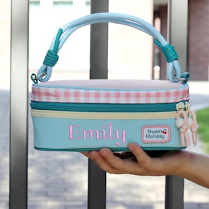 Op de afbeelding: Een lichtblauw rechthoekig etui met een roze en wit ruitjespatroon, een groenblauwe rits en een lichtblauw handvat. De naam Emily is in het roze op de voorzijde geborduurd, naast een rechthoekig label met de tekst Sweet Holiday.