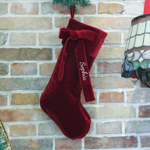 May include: A deep red velvet Christmas stocking featuring a matching velvet bow at the top. The stocking has a long, hanging ribbon tail embroidered with the name Sophia in white cursive script, centered on the front of the fabric.