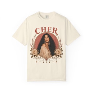 Może przedstawiać: Kremowy t-shirt z krótkim rękawem z nadrukiem graficznym przedstawiającym Cher. Wzór zawiera napis CHER czerwonymi literami nad okrągłym portretem artystki, otoczonym kwiatowymi ilustracjami, wiszącymi koralikami i różami.