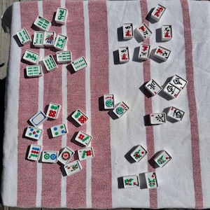 May include: A collection of white rectangular Mahjong tiles featuring various green, red, and blue engraved symbols, characters, and numbers. The tiles are scattered across a white fabric surface with prominent vertical red stripes.