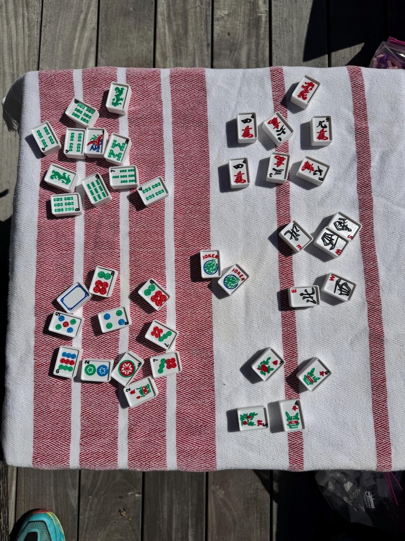 May include: A collection of white rectangular Mahjong tiles featuring various green, red, and blue engraved symbols, characters, and numbers. The tiles are scattered across a white fabric surface with prominent vertical red stripes.