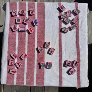 May include: A collection of rectangular pink mahjong tiles with various colorful symbols, numbers, and characters printed on their surfaces. The tiles are scattered across a white and red striped fabric surface, displaying diverse game markings.