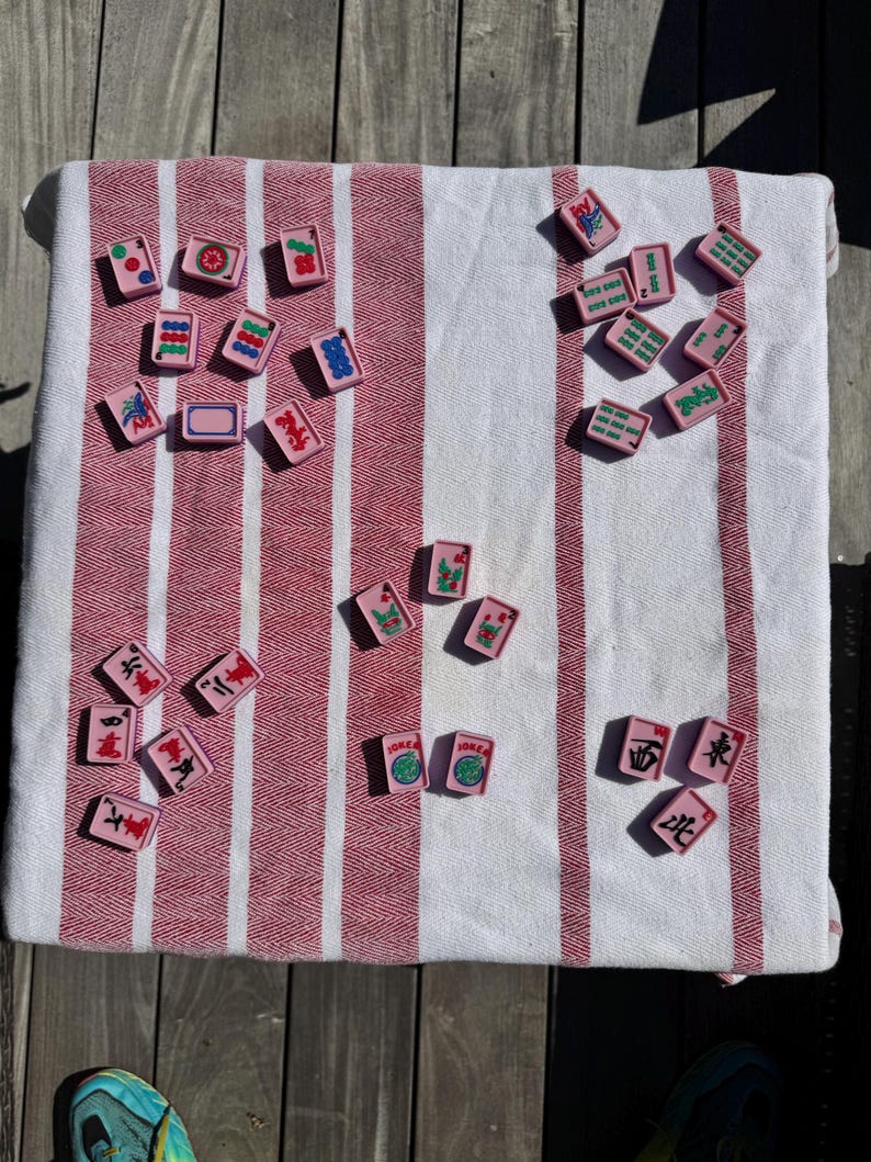 May include: A collection of rectangular pink mahjong tiles with various colorful symbols, numbers, and characters printed on their surfaces. The tiles are scattered across a white and red striped fabric surface, displaying diverse game markings.