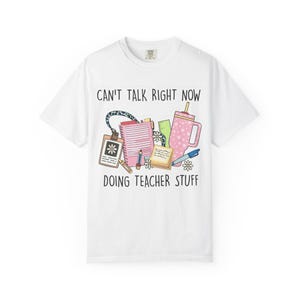 Puede incluir: Una camiseta blanca de manga corta con un estampado gráfico de material escolar que incluye un cordón, cuaderno, regla, notas adhesivas, bolígrafo y un vaso. El texto dice CAN'T TALK RIGHT NOW DOING TEACHER STUFF, rodeado de pequeñas flores blancas.