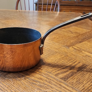 May include: A round copper saucepan with a polished metallic exterior and a dark, matte interior. The cookware features a long, slender, dark-colored metal handle attached to the side with a sturdy, visible rivet, extending outward from the main body.