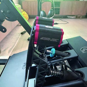 May include: A black and magenta Simagic racing pedal set featuring a metal frame, exposed springs, and a teal adjustment knob. The unit includes a black base plate with the word PRO printed on it, connected by black cables to the pedal assembly.
