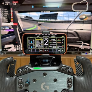 May include: A black racing steering wheel with carbon fiber paddle shifters and a central smartphone mount displaying a digital telemetry dashboard with speed, RPM, and tire temperature data, positioned in front of a large screen showing a race track.