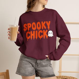 以下が含まれることがあります： オレンジ色の文字で「SPOOKY CHICK」と滴るようなフォントで書かれ、笑顔の小さな白い漫画の幽霊のグラフィックが添えられた、マルーン色の長袖クルーネックのスウェットシャツ。グレーのデニムショートパンツと合わせて着用。