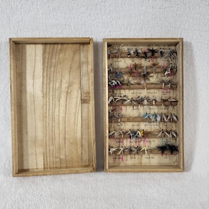 May include: A rectangular wooden box with a separate lid, containing a collection of fishing flies arranged in rows. The interior of the box features printed labels beneath each fly, showcasing various feather textures and colors like black, white, and pink.