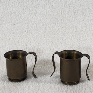 May include: Two small metallic vessels with a dark, tarnished brass finish. The vessel on the left features a single curved handle and a small pouring spout, while the vessel on the right features two symmetrical curved handles on opposite sides.