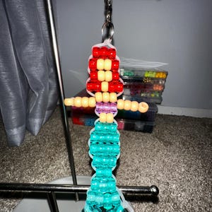 May include: A mermaid-shaped keychain crafted from colorful plastic pony beads strung on clear elastic cord. The figure features bright red hair, a peach-colored torso, a purple midsection, and a long, shimmering turquoise tail with a flared fin.