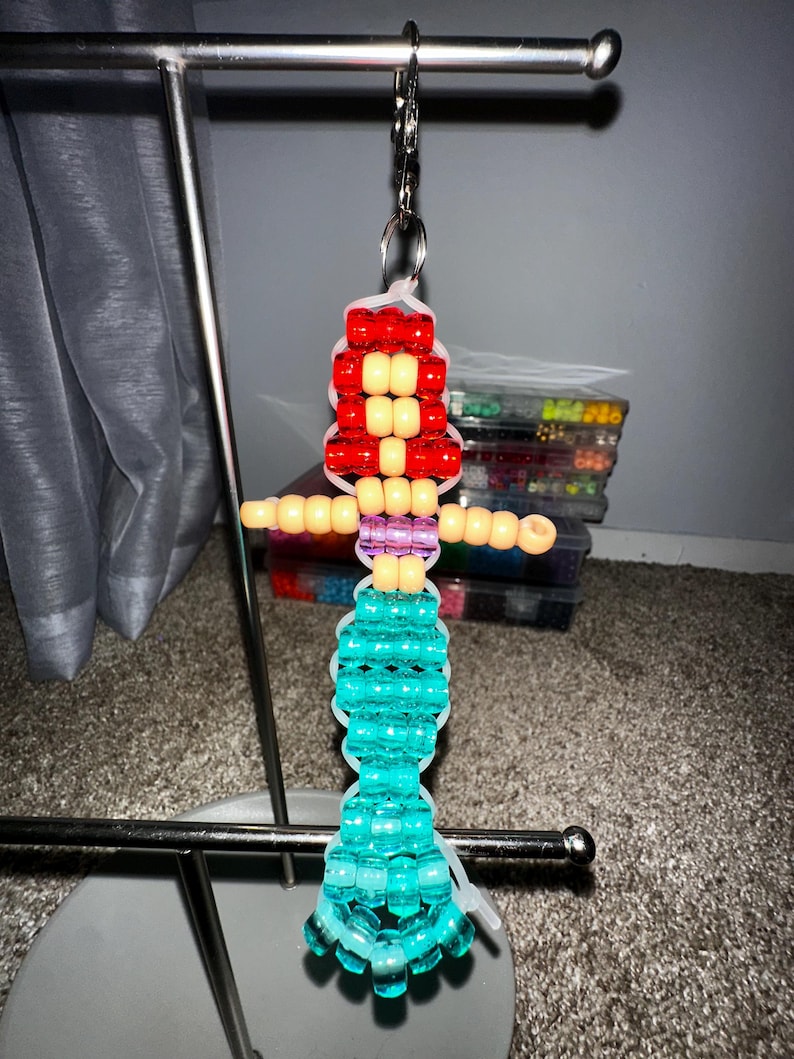 May include: A mermaid-shaped keychain crafted from colorful plastic pony beads strung on clear elastic cord. The figure features bright red hair, a peach-colored torso, a purple midsection, and a long, shimmering turquoise tail with a flared fin.