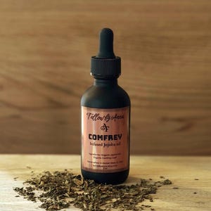 May include: A matte black glass dropper bottle labeled Tallow by Anna Comfrey Infused Jojoba oil, containing 2 oz of product. The label lists organic jojoba oil and organic comfrey leaf as ingredients. Dried green comfrey leaves are scattered below.