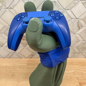 May include: A matte olive green sculpted hand stand featuring a blue wristband, holding a bright blue wireless video game controller. The controller has standard directional buttons, two analog sticks, and the classic triangle, circle, cross, and square symbols.