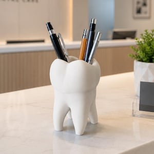 Peut inclure: Un porte-stylo blanc en forme de dent contenant plusieurs stylos, posé sur un comptoir en marbre. À côté se trouvent une petite plante en pot et une carte de visite noire avec le texte SMILE DENTAL CARE et une icône de dent imprimés en blanc.