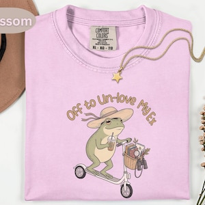 Può includere: Una t-shirt rosa chiaro con la grafica di una rana verde che indossa un cappello da sole, guida un monopattino e tiene una bevanda. Il cestino sul monopattino contiene libri e binocoli. Il testo sopra la rana recita, Off to Un-love My Ex.