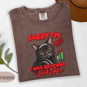 Puede incluir: Una camiseta color espresso con el gráfico de un gato negro que lleva gafas de estampado de leopardo y una diadema de cuentas que dice BCBA. El texto SORRY I'M LATE I WAS GRAPHING DATA rodea un gráfico de barras de colores con una flecha.