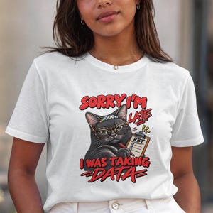Könnte beinhalten: Ein weißes kurzärmeliges T-Shirt mit einer Grafik einer schwarzen Katze, die eine Brille und ein Perlenstirnband trägt. Die Katze hält ein Klemmbrett mit der Aufschrift RBT und einen roten Stift. Der Text lautet SORRY I'M LATE I WAS TAKING DATA.