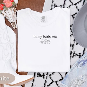 Può includere: Una t-shirt bianca a maniche corte con il testo ricamato in nero 'in my bcaba era' centrato sul petto, accompagnato da una piccola grafica di tre bambini stilizzati e due minuscoli cuori posizionati direttamente sotto il testo.