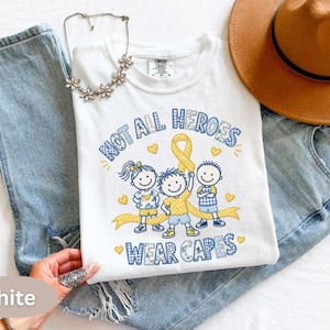 May include: A white t-shirt featuring a graphic of three children and a yellow awareness ribbon. The text reads Not All Heroes Wear Capes in blue and white checkered lettering. The design includes small yellow hearts and blue accents.