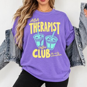 Könnte beinhalten: Ein violettes Kurzarm-T-Shirt mit gelbem und hellblauem Grafiktext, der ABA THERAPIST COFFEE CLUB the early intervention lautet, begleitet von Abbildungen zweier Eiskaffeebecher mit Strohhalmen auf der Vorderseite.