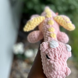 May include: A handmade crochet dragon plush toy featuring a soft pink body with a textured yarn finish. The creature has pale yellow wings and a matching yellow spine ridge, along with small white accents on its head and dark, oval-shaped eyes.