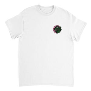 以下が含まれることがあります： 左胸に小さな円形のグラフィックが施された白い半袖Tシャツ。グラフィックには、黒い円形の枠の中に、鋭い歯と白い目を持つ緑色の質感のあるモンスターの頭が描かれており、背景は無地のピンク色になっています。