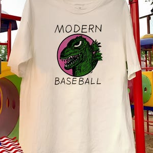 Op de afbeelding: Een wit t-shirt met korte mouwen en een grafische print van een groene monsterkop in een roze cirkel. De tekst "MODERN" staat in het zwart boven de cirkel gedrukt en de tekst "BASEBALL" staat in het zwart onder de cirkel.