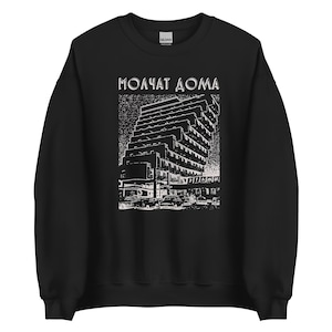 Peut inclure: Un sweat-shirt noir à col rond avec une impression graphique blanche représentant un bâtiment brutaliste et des voitures. Le texte "МОЛЧАТ ДОМА" est imprimé au-dessus du bâtiment et le mot "ЭТАЖИ" est visible sur la partie inférieure du bâtiment.