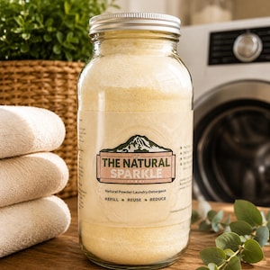 May include: A clear glass jar filled with pale yellow laundry detergent powder, topped with a silver metal lid. The label features a mountain graphic and the text THE NATURAL SPARKLE, Natural Powder Laundry Detergent, REFILL, REUSE, REDUCE.