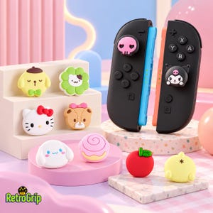 May include: Two black game controllers with blue and orange side rails, featuring character-themed thumbstick covers. Several silicone thumbstick caps shaped like cartoon animals, flowers, and food items are displayed on tiered platforms.