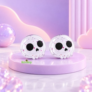 Puede incluir: Dos agarres circulares de resina transparente con un diseño de calavera, ojos redondos negros y una pequeña nariz negra. El material translúcido contiene destellos de purpurina rosa y morada. El texto RetroGrip está impreso en verde.