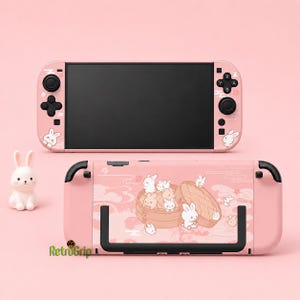 May include: A light pink handheld gaming console featuring a screen and controllers with a white rabbit graphic. Below, a matching console backplate displays a bamboo steamer basket filled with small white and tan rabbits, alongside a small rabbit figurine.