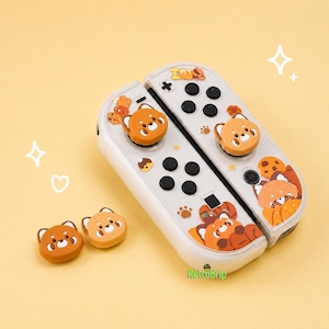Puede incluir: Un par de mandos de juego blancos con gráficos de panda rojo naranja y fundas para joystick a juego. Los mandos incluyen botones negros, una cruceta y pequeños detalles de huellas. El texto RetroGrip es visible en la imagen.