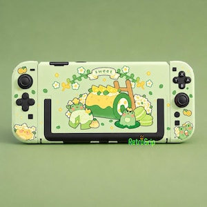 Op de afbeelding: Een lichtgroene spelcomputer met een skin voorzien van een illustratie van een groene kikker, een gele rolcake en macarons. Het ontwerp bevat witte bloemen, citroenschijfjes en de tekst sweet. Zwarte knoppen en joysticks zijn zichtbaar.