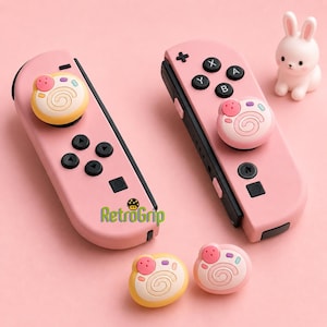 May include: Two pink game controllers featuring black buttons and custom thumbstick caps shaped like swirled pastries with pink, yellow, and blue accents. A small white rabbit figurine sits nearby, and the text RetroGrip is printed in green.