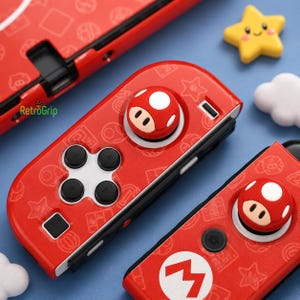 Peut inclure: Une manette de jeu vidéo rouge avec une croix directionnelle blanche, un capuchon de joystick en forme de champignon et un logo M blanc. La surface est ornée d'un motif répétitif de petites icônes incluant des étoiles, des pièces et des champignons.