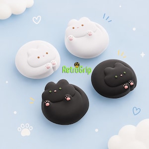 May include: Four circular silicone grips labeled RetroGrip, featuring two white and two black cat designs. Each cat has small ears, a tiny nose, and raised pink paw pads on its front paws. The smooth, matte-finish grips are arranged on a light blue background.